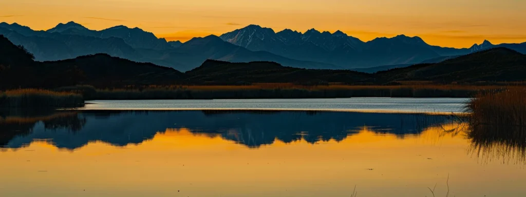 a vibrant sunset casts a golden hue over a serene lake, reflecting the majestic silhouettes of towering mountains in the tranquil water below.