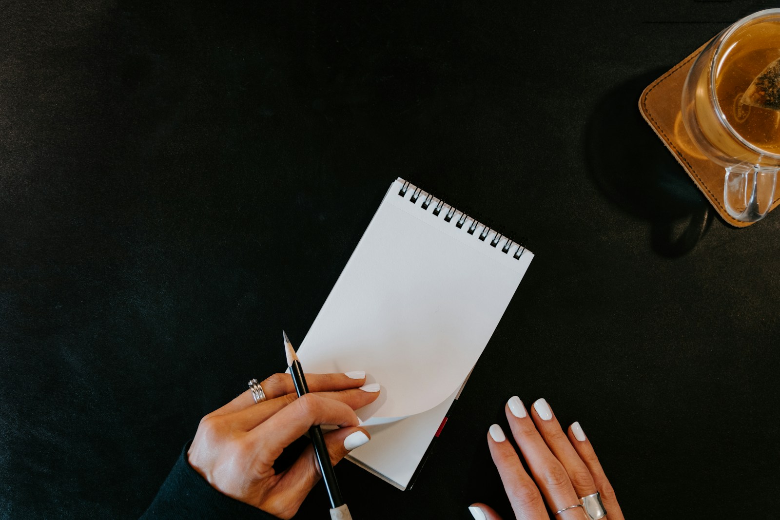 Hands writing in a small notebook with a cup of tea on a coaster nearby