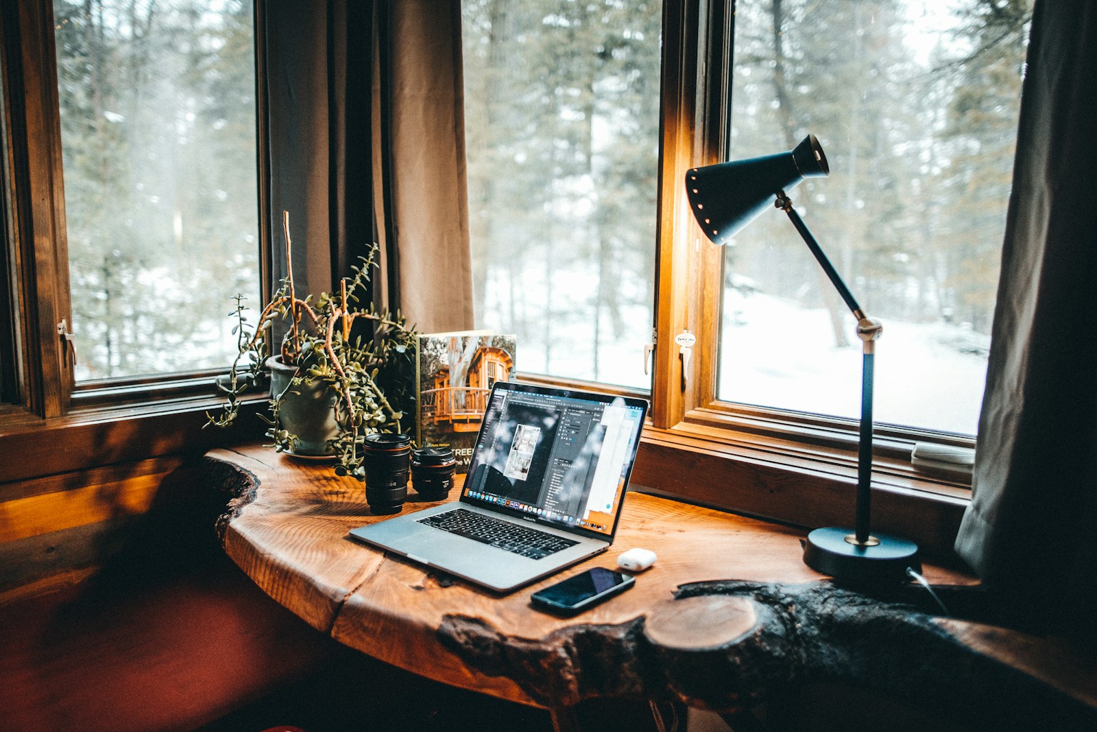 An open laptop on a rustic wooden desk by a large window with snowy trees outside, warm lamp light