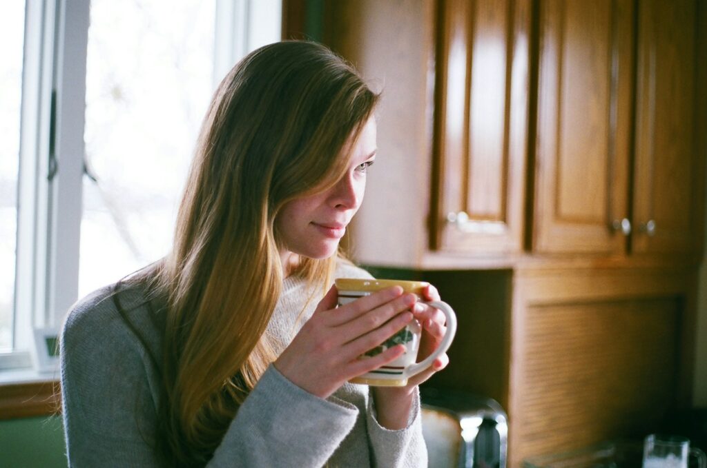 An adult in casual clothing holding a ceramic mug with both hands at a kitchen table in soft natural light