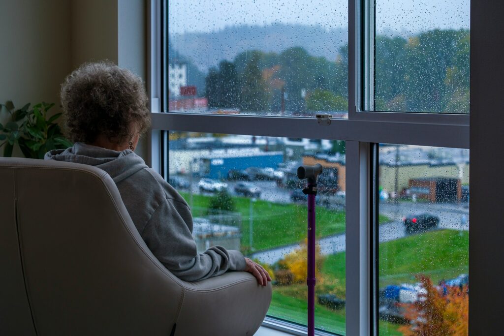 An adult sitting in a chair by a large window looking outside at a rainy day with reflective contemplative mood