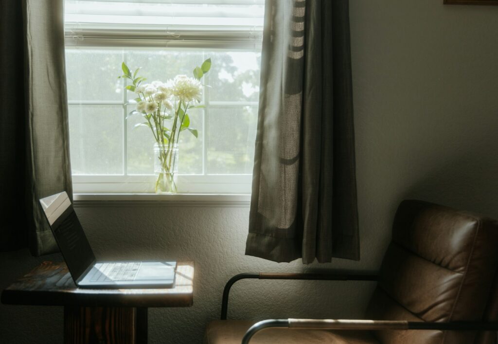 An open laptop and small bouquet of flowers on a desk by a sunlit window with a chair