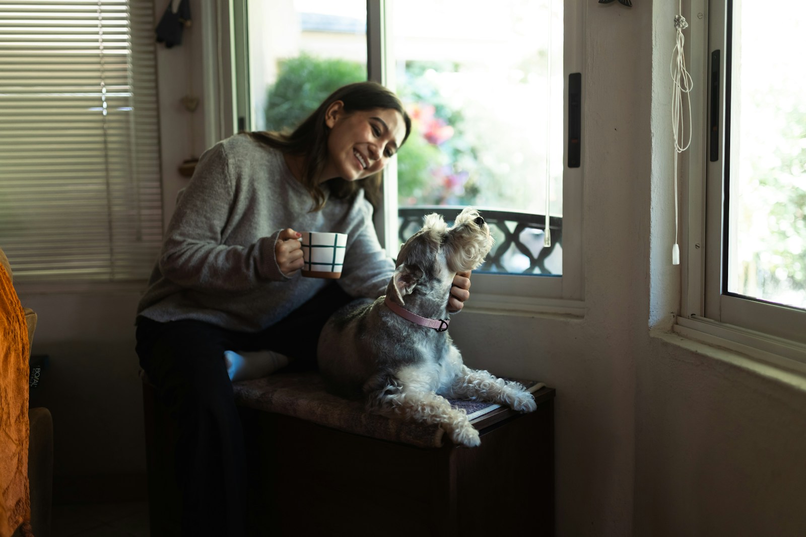 A woman holding a coffee cup while petting a dog by a window