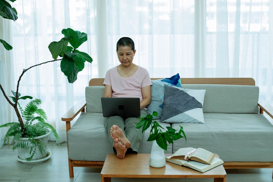 Full body of serious mature Asian lady in casual clothes sitting on couch with pillows and browsing on netbook next to table with books and window with curtains in apartment