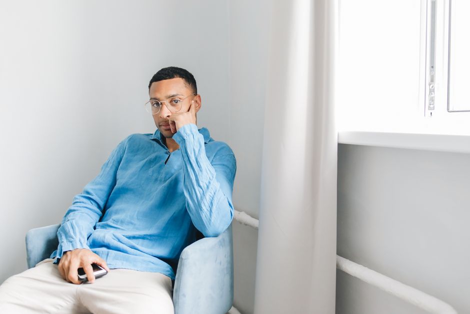 Man in a blue long-sleeved shirt sitting on a chair near a window, looking thoughtful