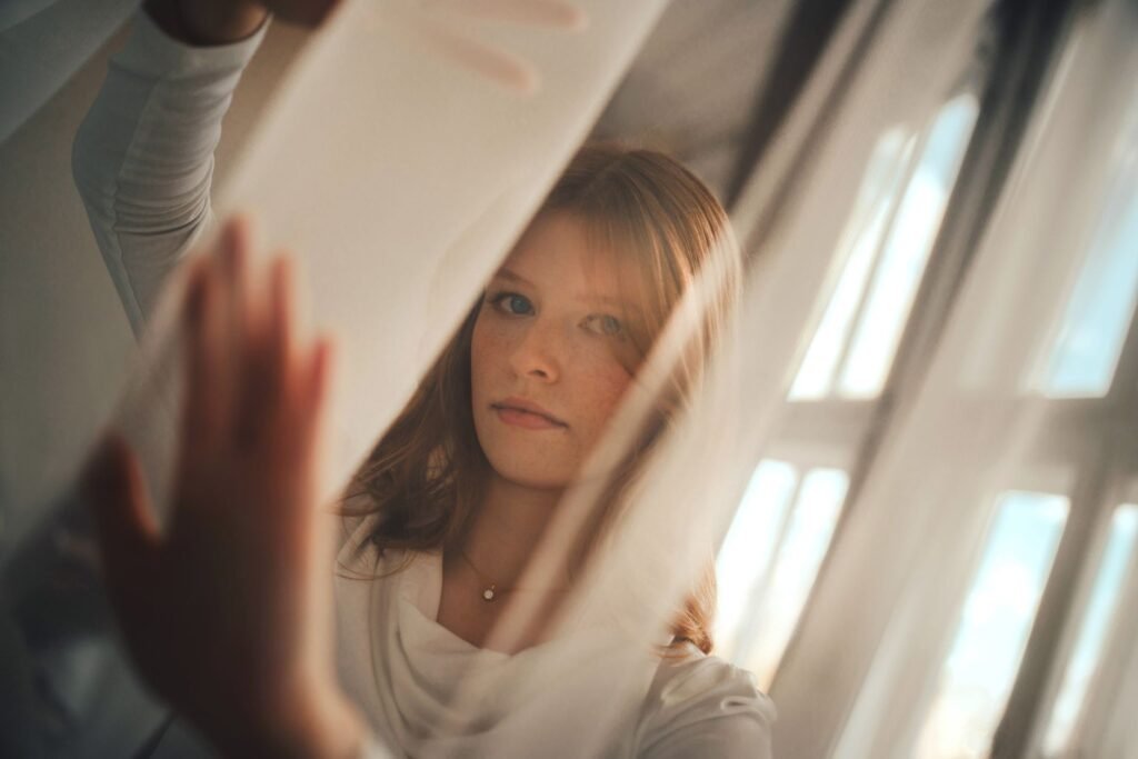 Portrait of a young woman partially obscured by sheer curtains with soft sunlight filtering through.