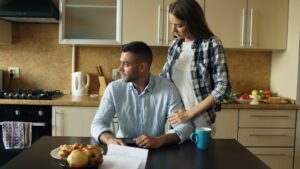 Upset young man reading unpaid bills and hugged by his wife supporting him early morning in the kitchen at home.