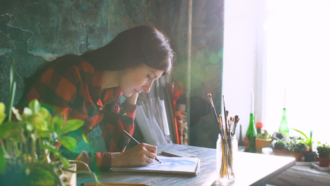 Young woman artist painting sketch on paper notebook with pencil in studio indoors. Flecks of sunlight.