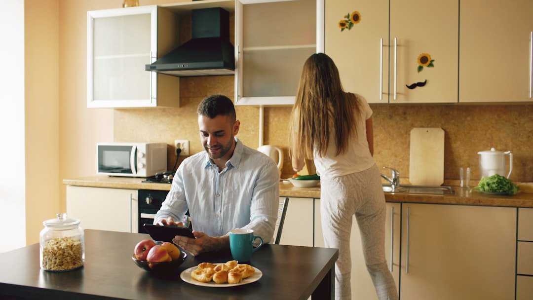 Happy young man using digital tablet computer sitting in the kitchen and talking his wife while she cooking at home in the morning.