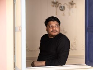 A man in a black sweater thoughtfully looks outside through a window. Captured indoors with soft lighting.