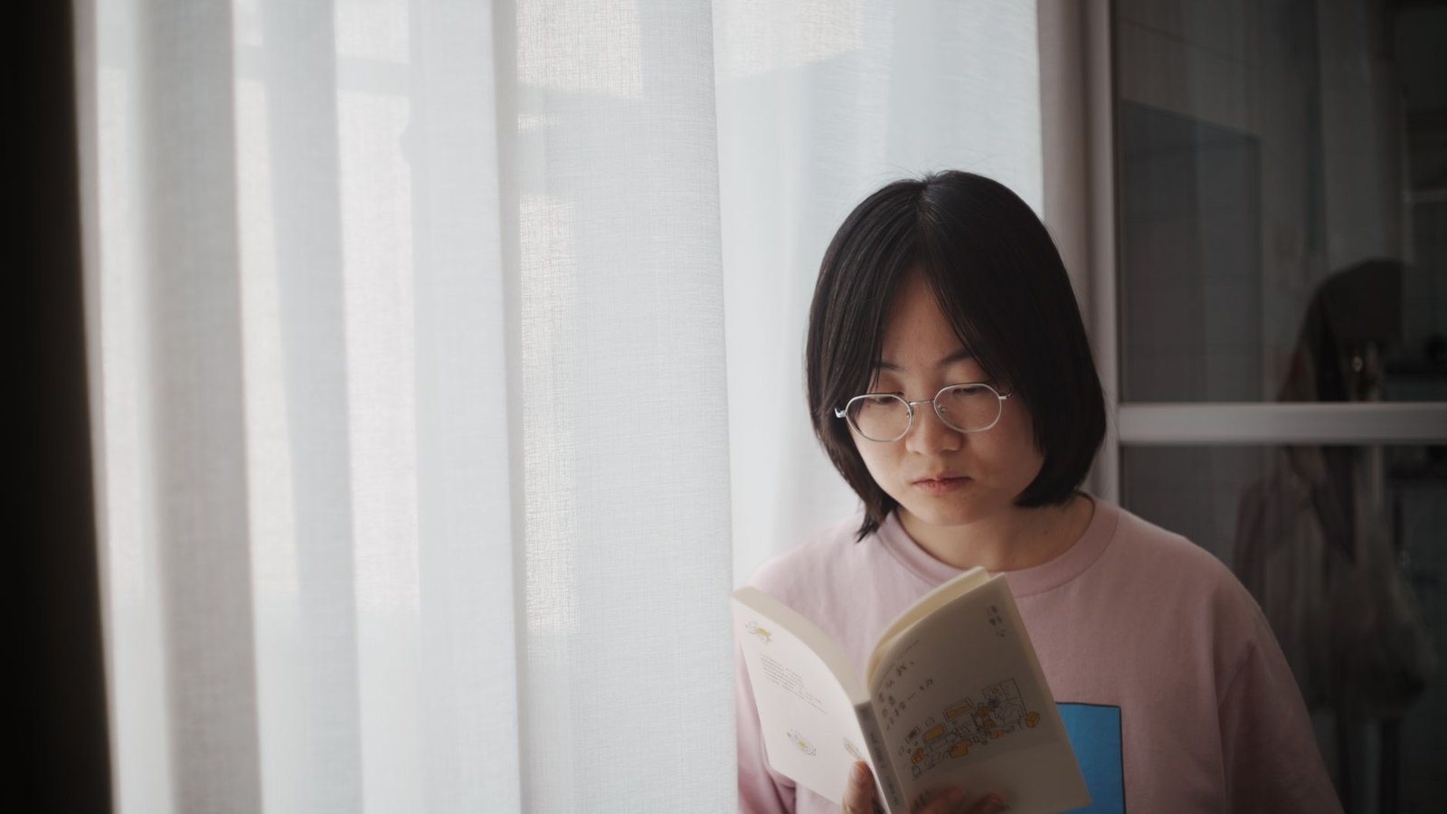 A young woman reads a book by a bright window, creating a calming indoor scene.
