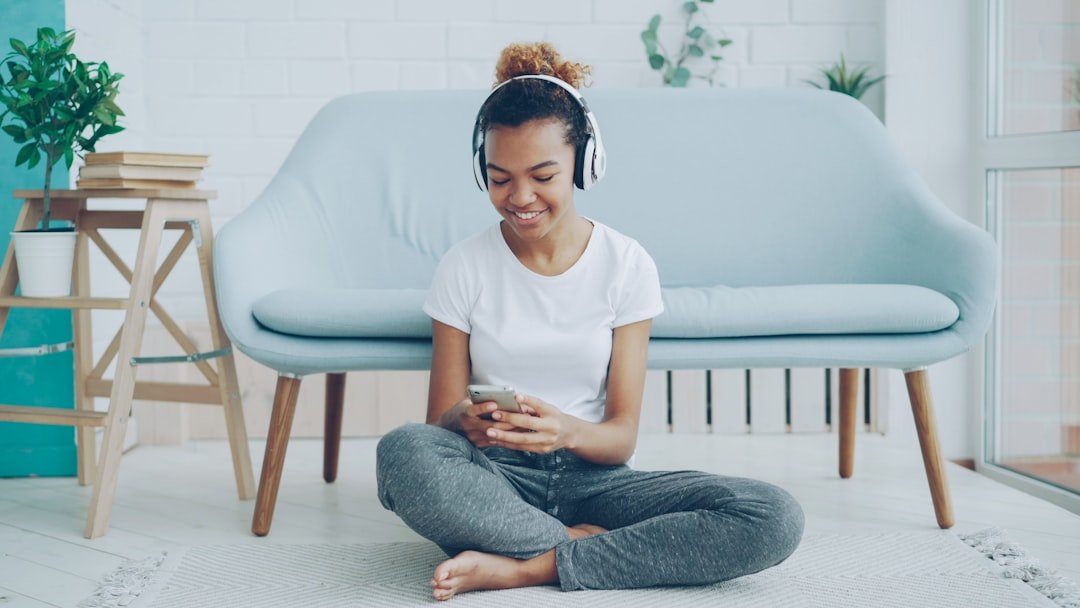 Pretty girl is playing game on smartphone touching screen and smiling while listening to music with headphones sitting on floor at home. Technology, millennials and fun concept.