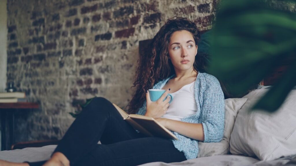 Cheerful girl is reading funny book in bedroom lying on comfy double bed, smiling and holding cup with drink. Hobby, happy people, beautiful interiors and literature concept.