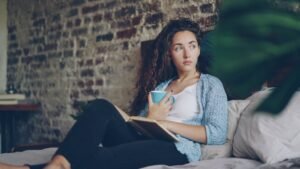 Cheerful girl is reading funny book in bedroom lying on comfy double bed, smiling and holding cup with drink. Hobby, happy people, beautiful interiors and literature concept.