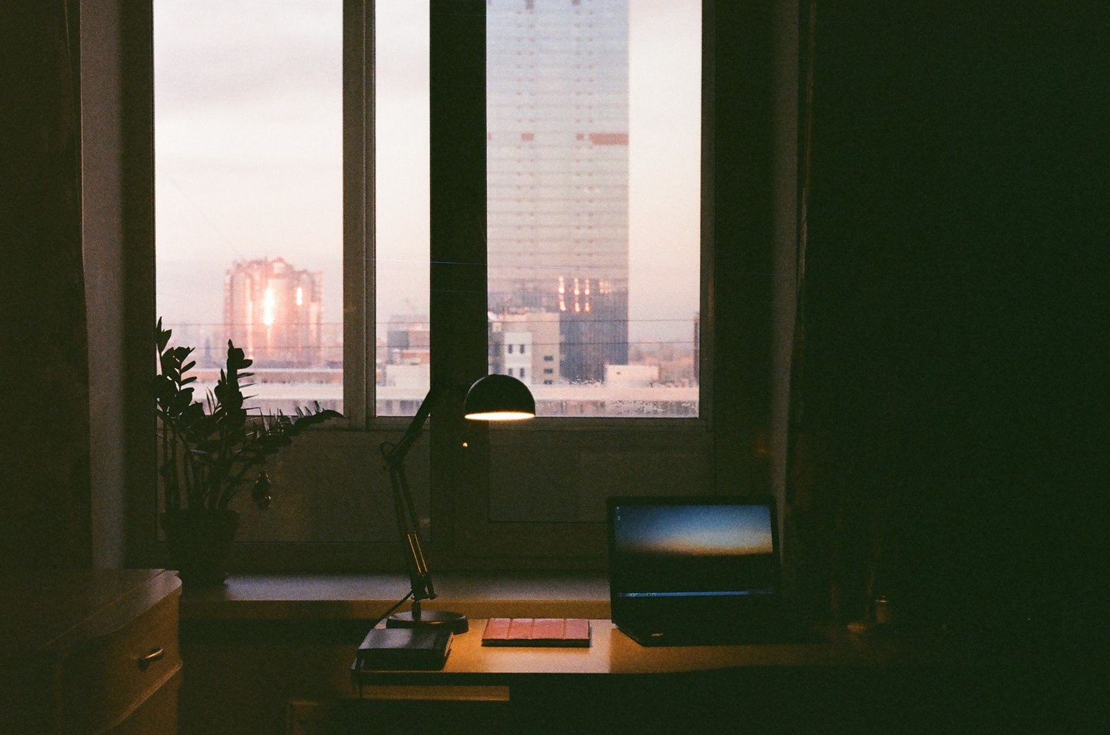 A cozy workspace with a laptop and lamp, overlooking a cityscape at dusk through a large window.
