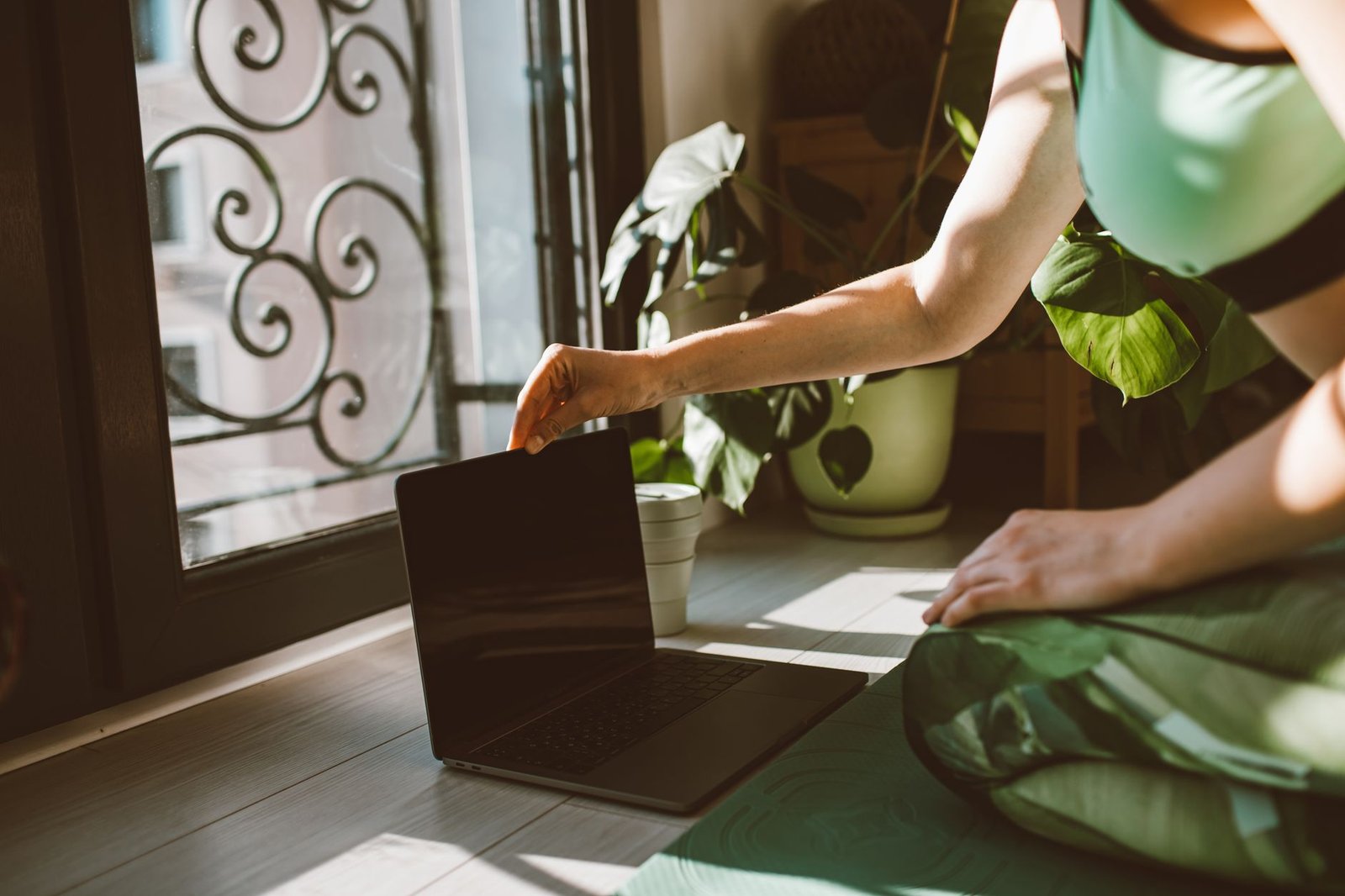 A woman sitting on a yoga mat using a laptop beside indoor plants, enjoying sunlight.