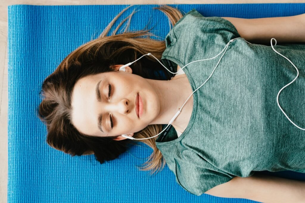 From above of calm young female in casual wear listening to music in earphones while taking break after workout on blue mat in flat