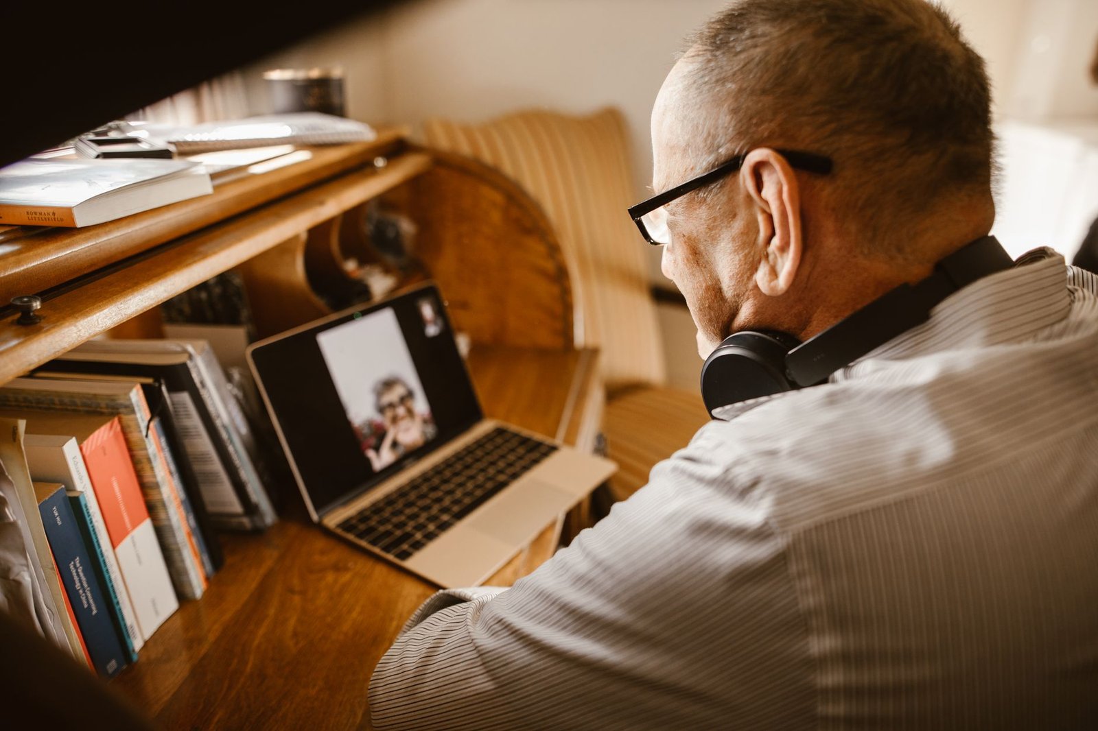 Senior man wearing headphones and eyeglasses on a video call using a laptop at a wooden desk.