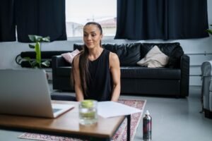 Woman sitting on floor using laptop, with a sofa and decor in cozy living room.
