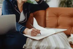 A woman with red nails writes in a notebook while sitting on a comfortable couch, using a laptop.