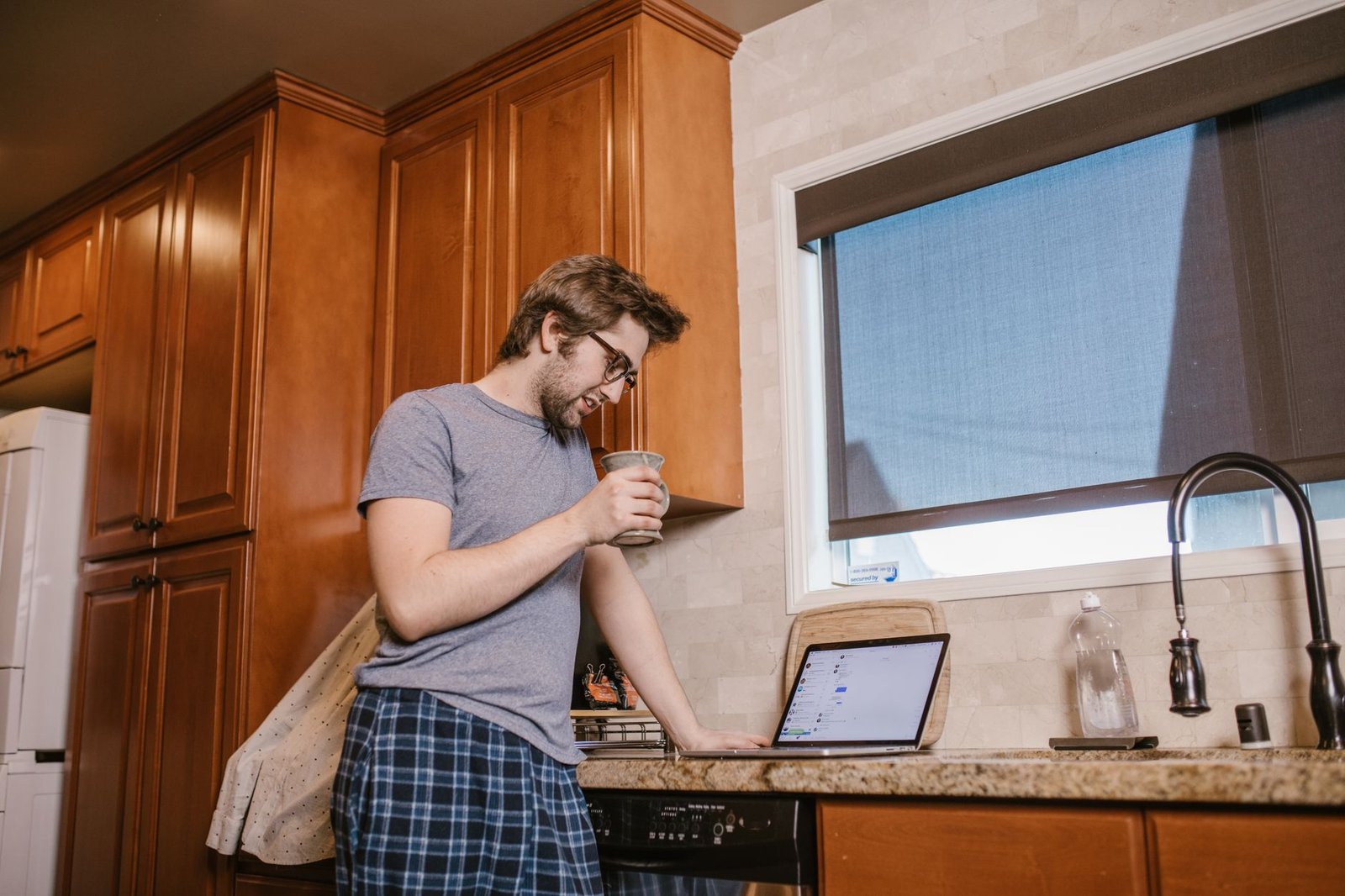 Man in pajamas enjoying coffee while browsing a laptop in a kitchen setting.