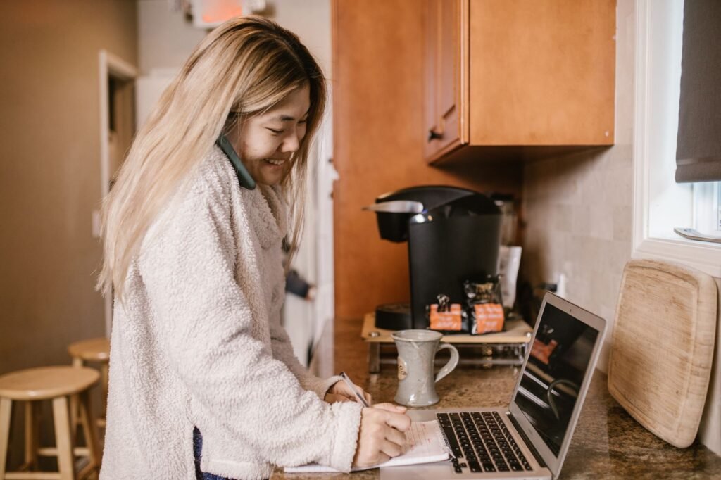 A woman multitasking at home, working on a laptop while on a phone call in a cozy kitchen setting.