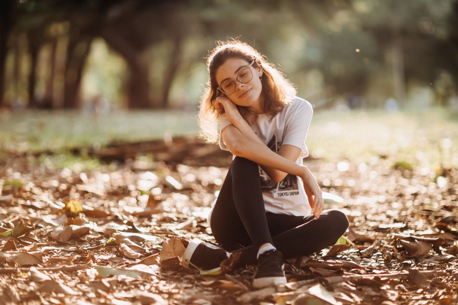 Young woman in glasses enjoys a tranquil moment outdoors among fallen leaves.