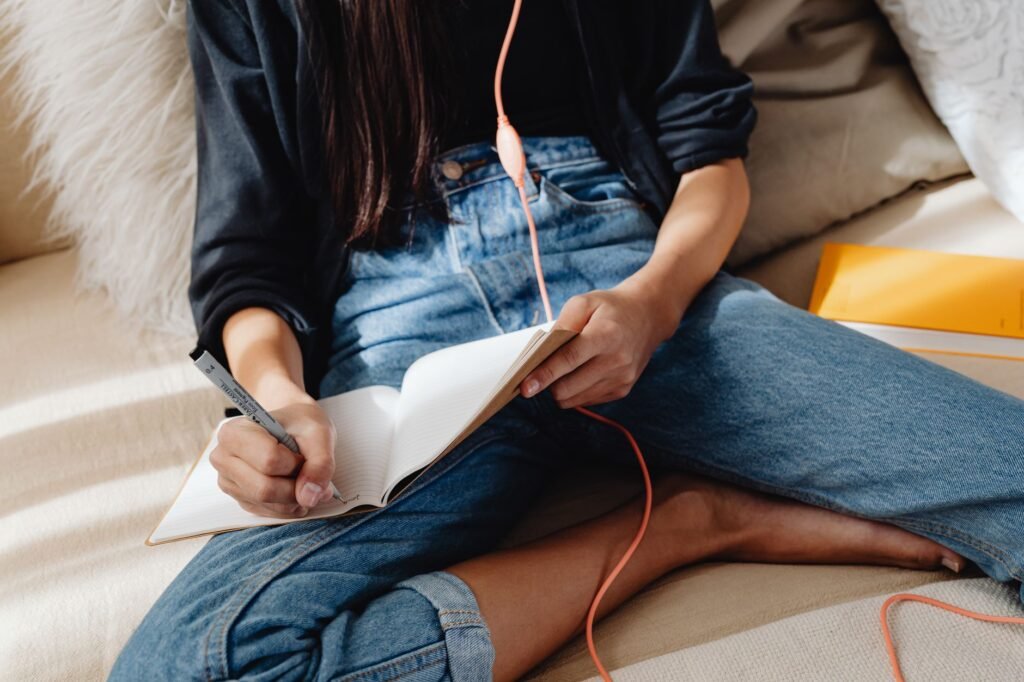 A young woman sits on a cozy couch, writing in a notebook, with earphones in. Casual and relaxed setting.