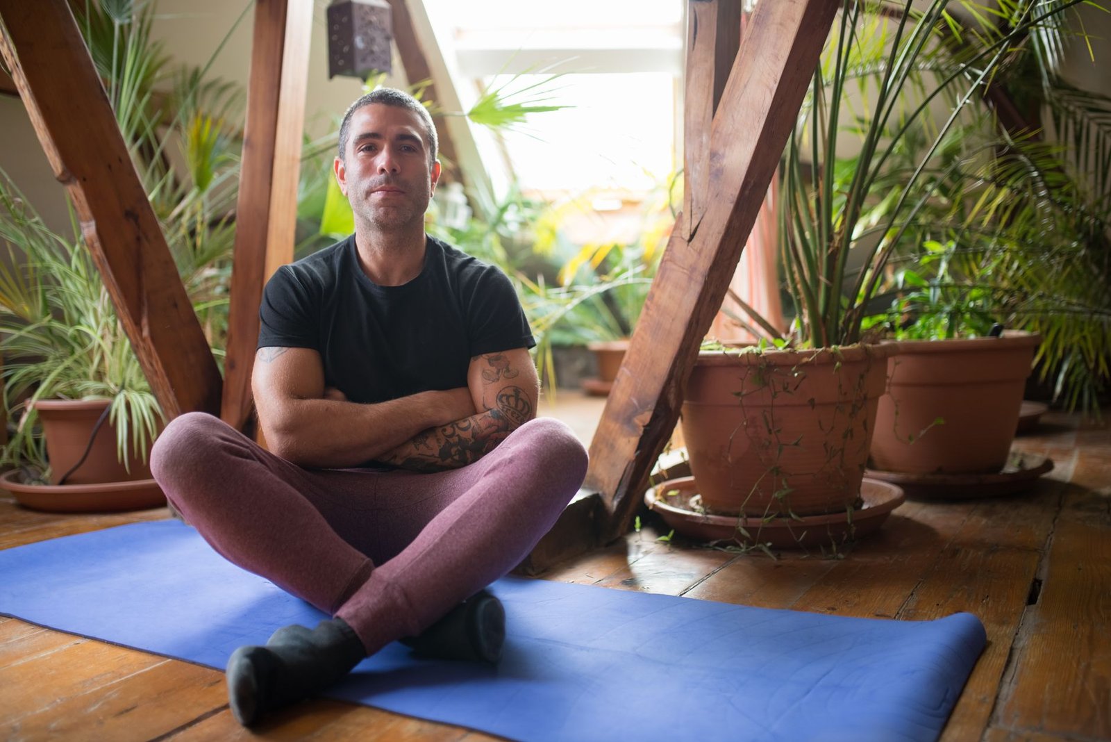 A man sits cross-legged on a yoga mat indoors, embracing a healthy lifestyle with surrounding greenery.