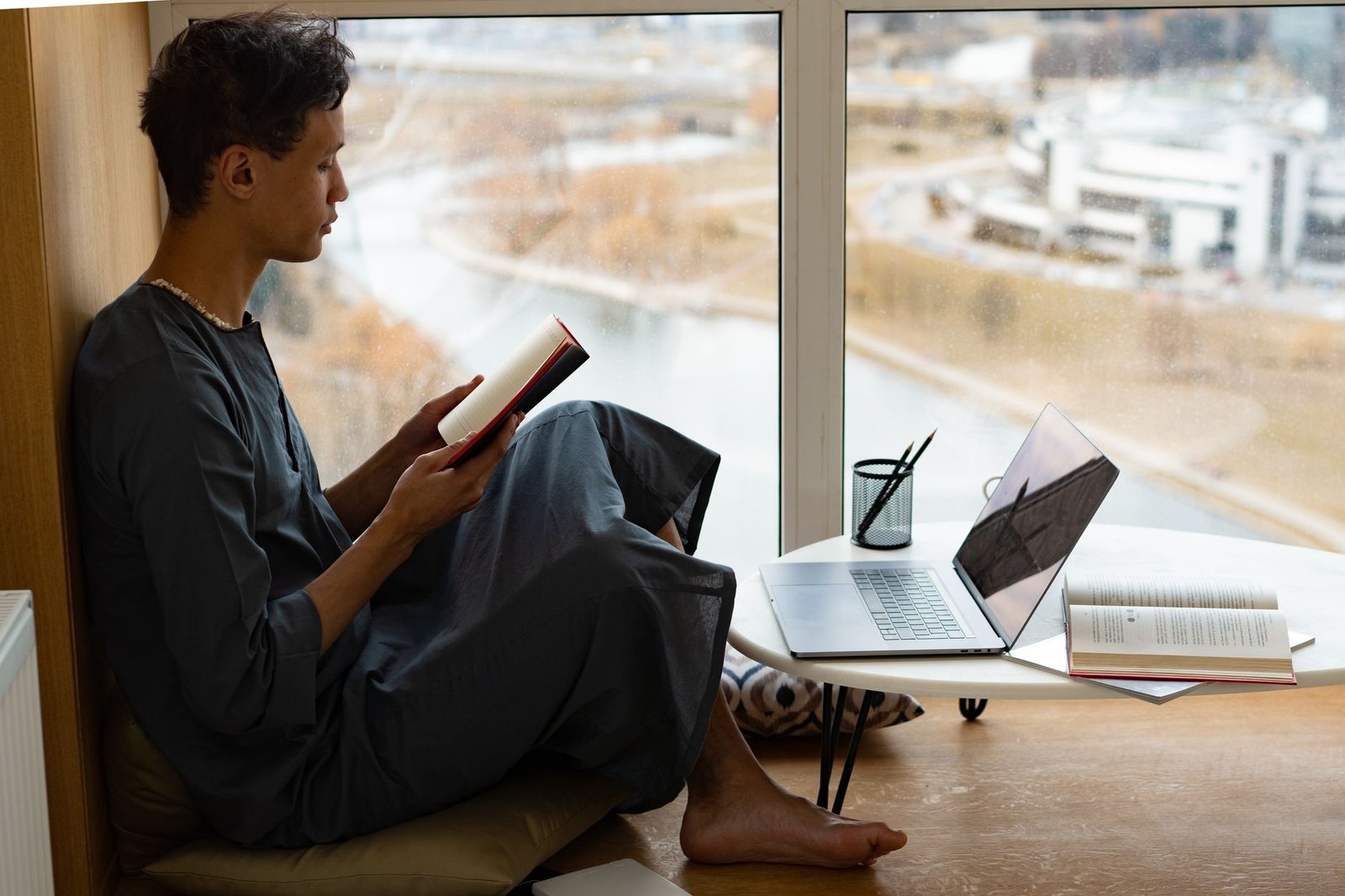 Adult man sits by a window reading a book next to a laptop on a table.