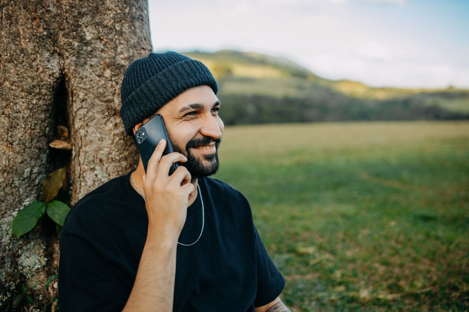 Smiling man with a beard talking on a smartphone against a rural outdoor backdrop.
