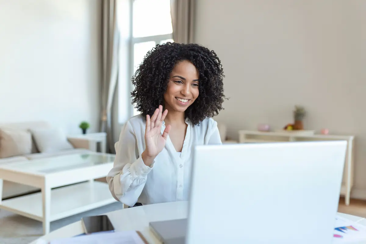 A woman waving hello to her laptop in a bright living room