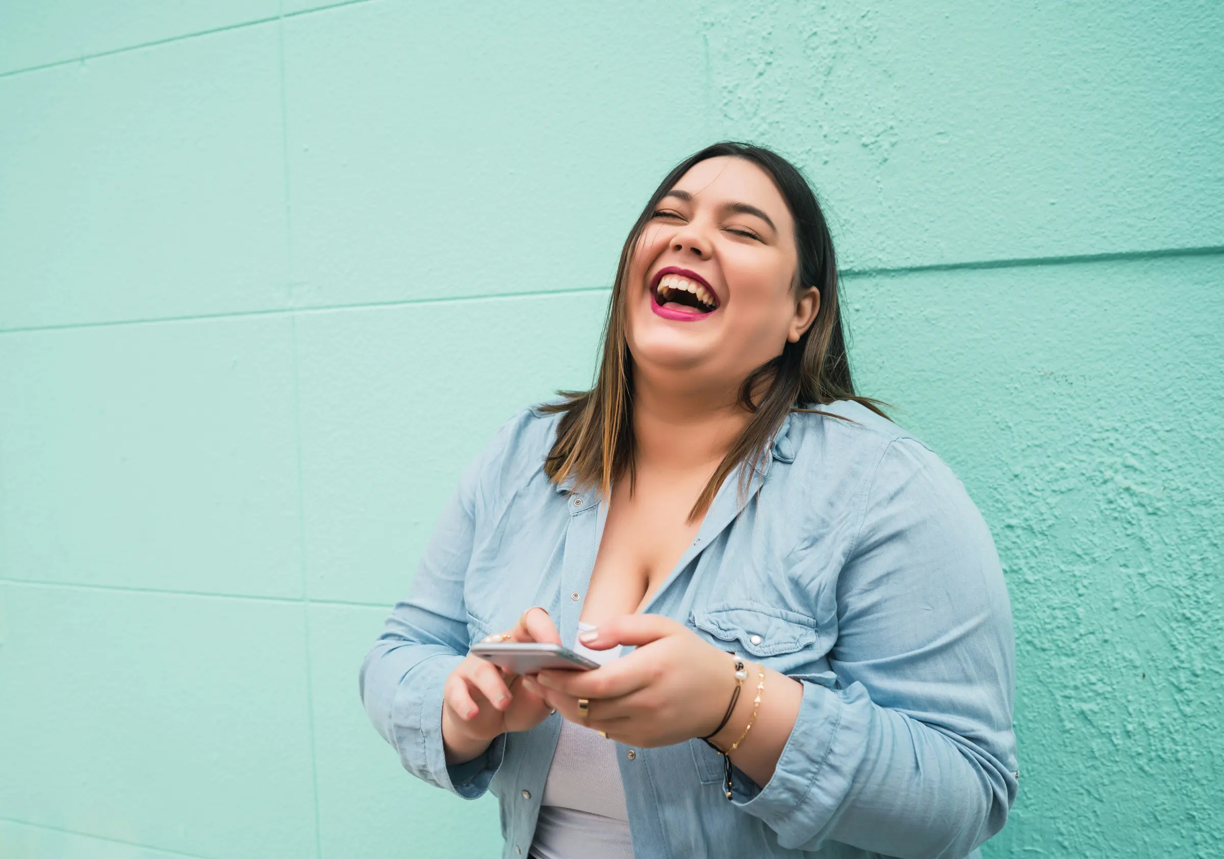 Woman laughing with her phone against a teal wall